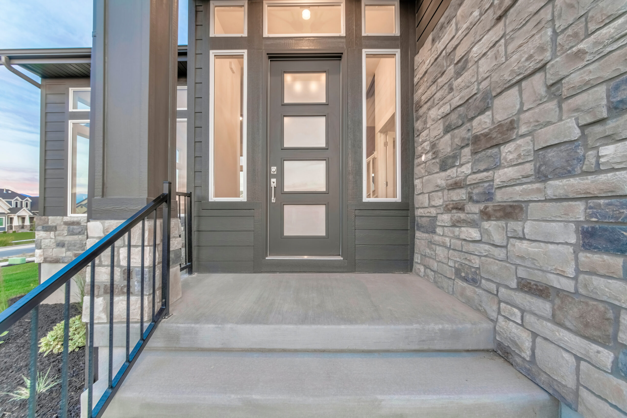 Front view of the house entrance with glass panelled front door and sidelights. Stairs with railing and gray stone brick wall can be seen at the entrance.