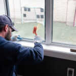 A bearded man wearing a cap and gloves works on a window frame with a mallet, overlooking a green lawn with a soccer net. Glacier_Glass_Regina