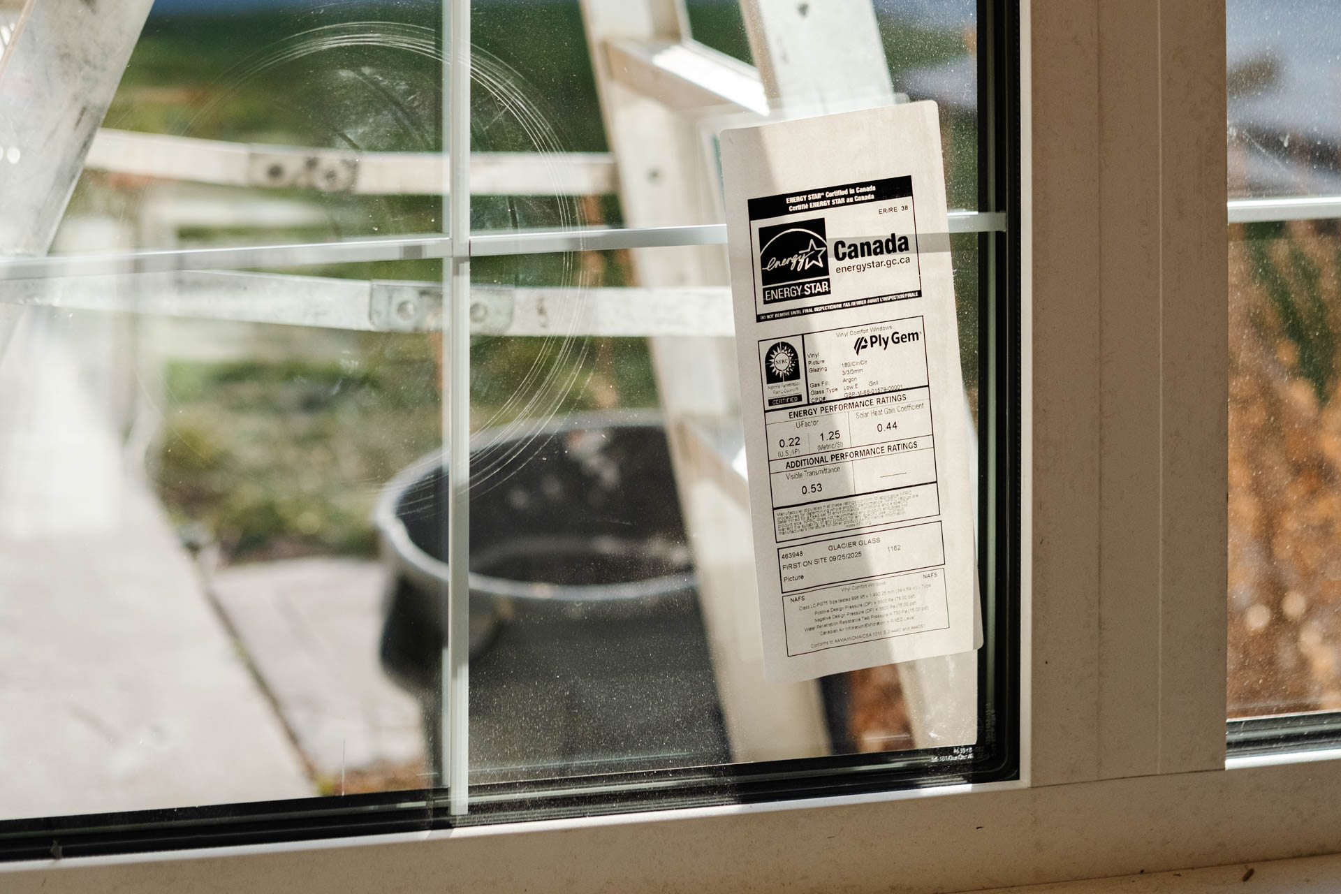 A dirty window with internal grilles displays an Energy Star label and reveals a blurry outdoor scene featuring a ladder and a dark bucket. Glacier_Glass_Regina