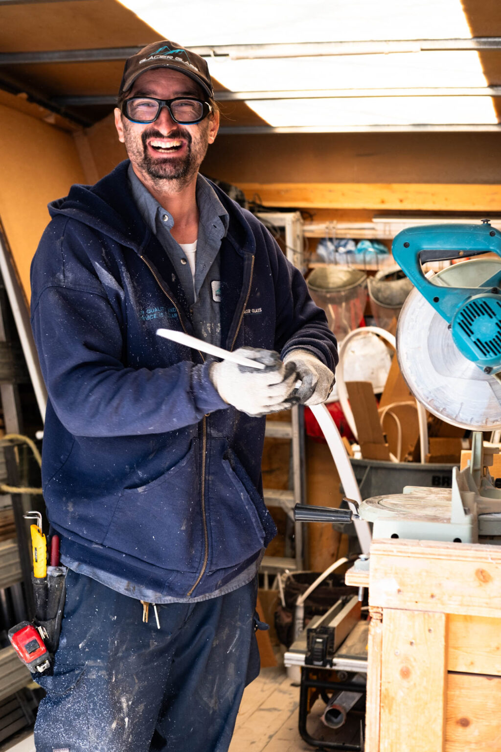 A bearded man in work clothes, smiling and wearing glasses and a cap, holds a white object while standing in a cluttered work area with a large teal saw nearby. Glacier_Glass_Regina