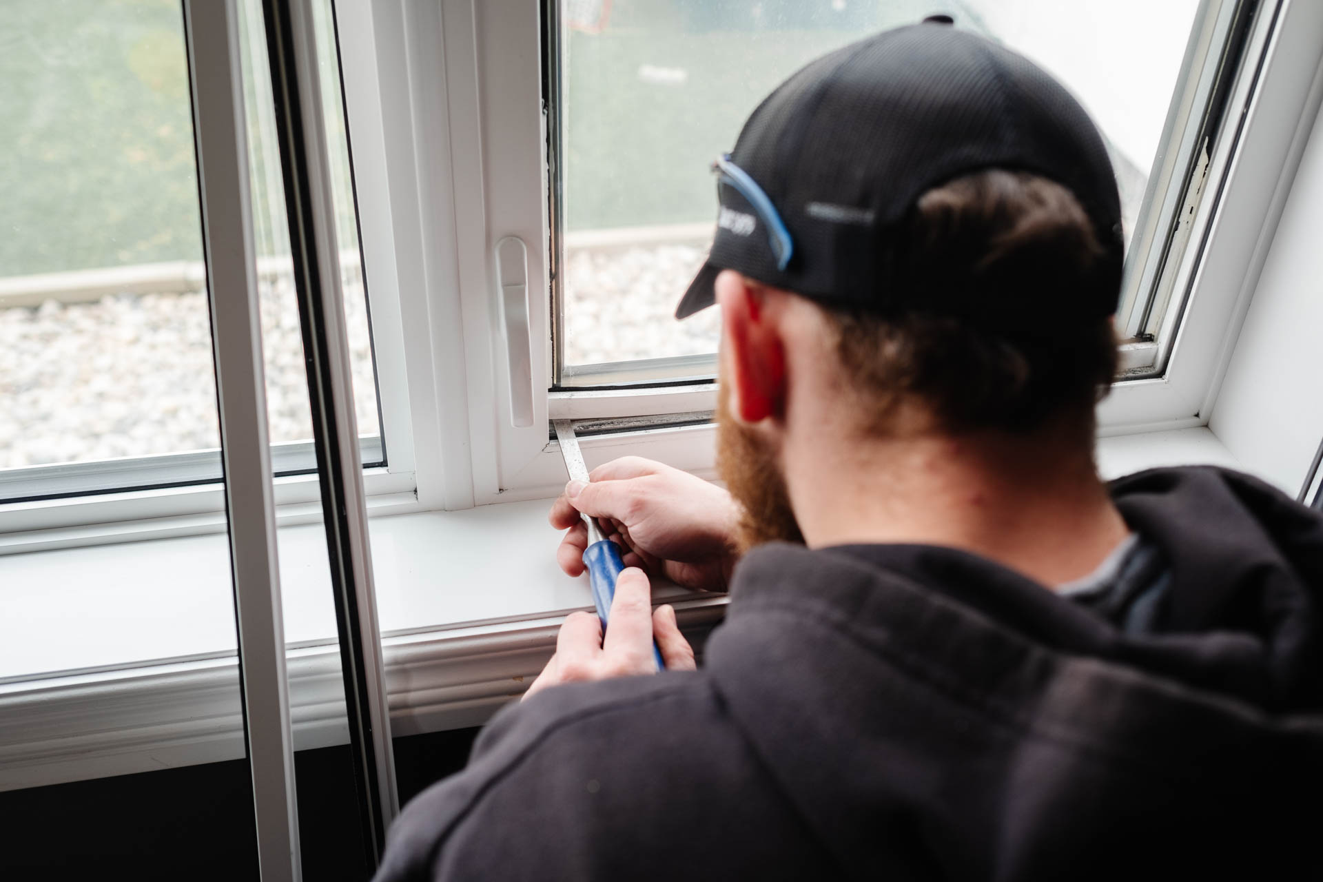 From behind, a bearded person in a dark hoodie and cap uses a tool on a white window frame, with gravel visible outside. Glacier_Glass_Regina